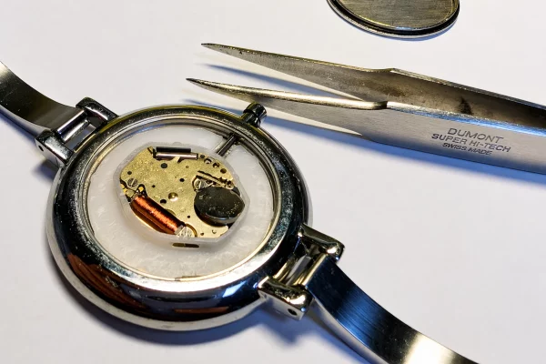 Otwarty zegarek kwarcowy z widocznym mechanizmem, obok pęseta zegarmistrzowska i zdjęta pokrywa koperty